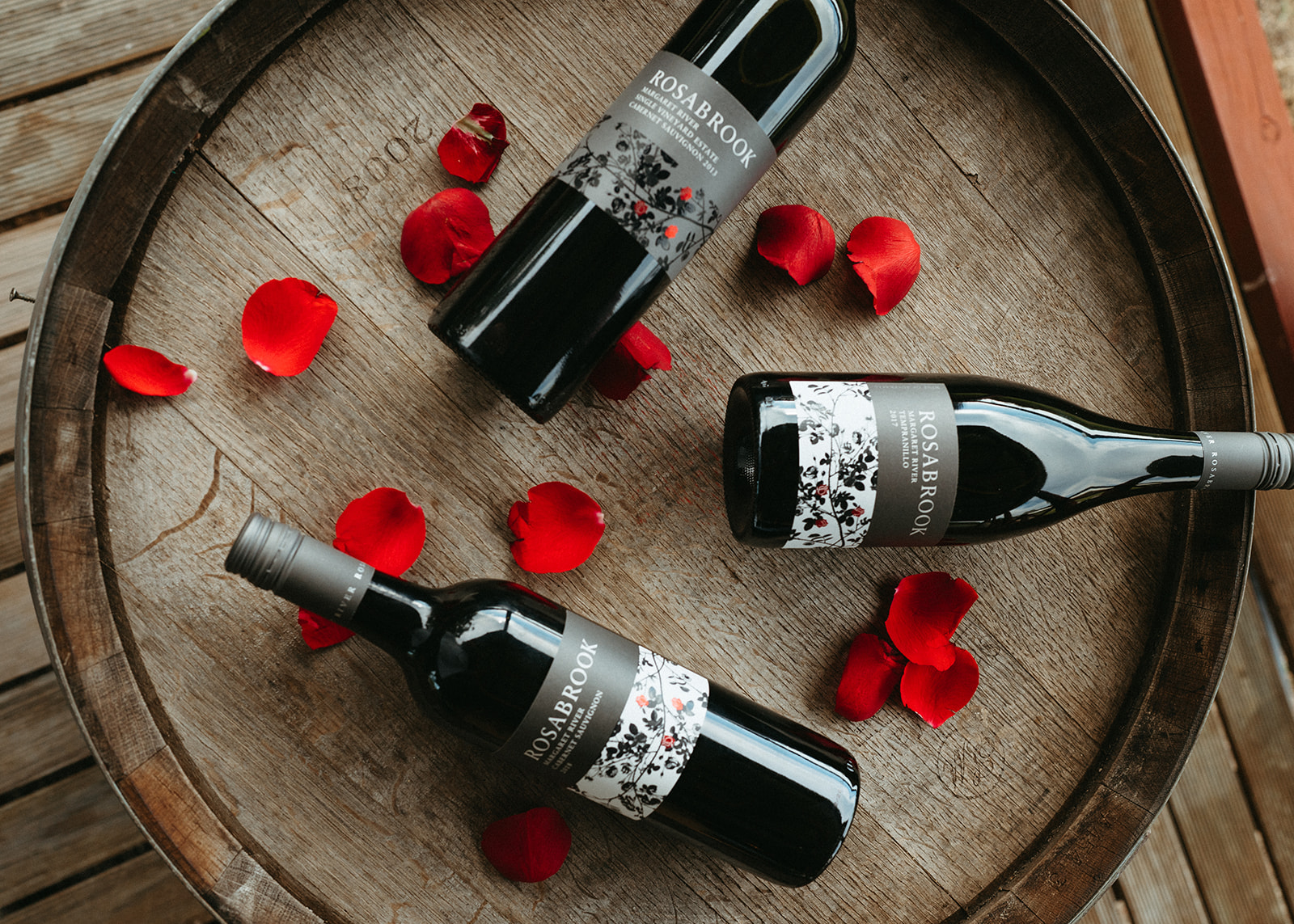 Three Rosabrook wine bottles on top of a barrel with rose petals 